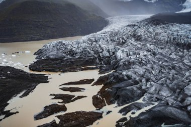 Hava görüntüsü, buzul diliyle Skaftafell, İzlanda 'da buzul kütleleri, koyu morina kanalları, sisli dağlar ve bulutlu aydınlık bulutlu bir gölle buluşan pürüzlü bir buzul dilini gösteriyor..