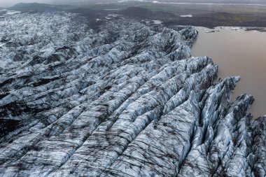 Hava görüntüsü, İzlanda 'da sütlü kahverengi bir göle doğru akan yarık bir buzulu gösteriyor. Mavi buz yumuşak bulutlu ışığın altında koyu kül ile çizilmiş ve düz tundraların ötesinde de düz tundralar var..