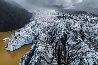 Vatnajokull, İzlanda 'daki sütlü kahverengi göllere doğru akan kül tabakası manzarası. Derin yarıklar, alçak bulutların altındaki mavi beyaz buzu geniş açılı bulutlu ışıkla keser..