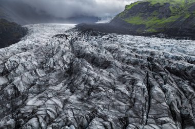 Hava görüntüsü volkanik tepeler ve İzlanda 'nın Vik kenti yakınlarındaki yosunlu yamaçlar arasında yarılmış bir buzulu gösteriyor. Kül çizgili buz, kasvetli bulutların altındaki bir vadiye doğru iniyor..
