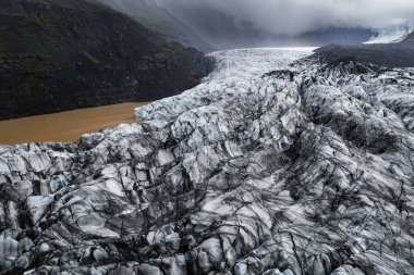 Havadan bakıldığında İzlanda 'da yarılmış bir Vatnajokull buzulu, koyu kül bantları, sivri dişli dişler ve düşük bulutların altında kalan sütlü kahverengi bir göl ve yayılmış ışık bulutu görülüyor..