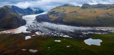 Havadan bakıldığında, İzlanda 'nın Vatnajokull bölgesindeki sütlü bir lagüne, yosunlu ovaları, volkanik yamaçları ve dağılmış gün ışığıyla koyu morfin çizgileri olan bir buzulun dili görülüyor..