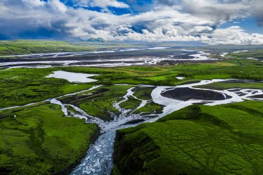 Landmannalaugar yakınlarındaki İzlanda dağlarında yosunlu düzlükler ve siyah kumlar boyunca örülmüş bir buzul nehrinin havadan görünüşü, bulutlar ayrılıyor ve gümüşi su gün ışığında..