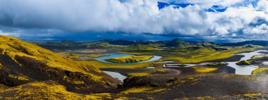 Turkuaz nehirler ve göller, Landmannalaugar yakınlarındaki İzlanda Dağları 'ndaki yosunlu lav tarlalarından ve kara kumlardan geçer. Rhyolite tepeleri ve fırtına bulutları yaz ışığı çerçevesinde.