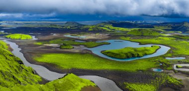 Landmannalaugar yakınlarındaki İzlanda volkanik dağlarının hava manzarası, yosun yeşil lav tarlaları kıvrımlı nehirleri ve düşük fırtına bulutlarının altındaki yansıtıcı gölleri çevrelemektedir..