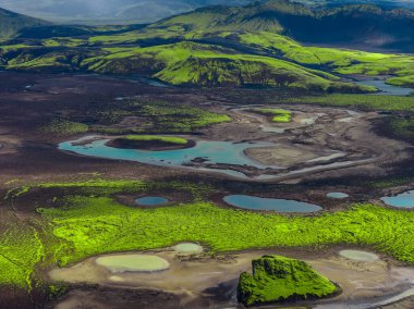 Karanlık lavlar, turkuaz göletler ve İzlanda Highlands 'taki krater jantları üzerindeki neon yosunlarının havadan görünüşü. Alçak tepeler, yumuşak ışık ve bulutların altında çember oluşturur..