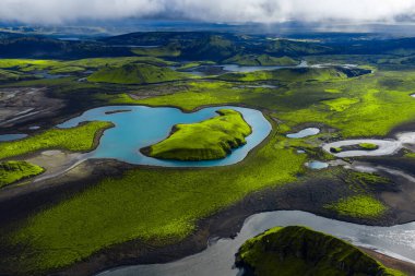 Hava manzarası, siyah volkanik kumlar içinde turkuaz bir göl içinde zümrüt bir ada ve İzlanda dağlık arazilerinde krater tepecikleri ve örülmüş dereleri olan yosunlu lav tarlaları gösteriyor..