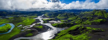 Hava manzarası, İzlanda 'da bazalt ve kül boyunca örülmüş bir buzul nehri, yosun yeşili yamaçlar, ryolit dağları ve yamalı bulutların altında gün ortası ışığı gösteriyor..