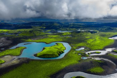Hava görüntüsü siyah lav üzerinde yosun, tepenin etrafında turkuaz bir göl, örülmüş nehirler ve Landmannalaugar yakınlarındaki İzlanda Dağları 'nda göletler gösteriyor..