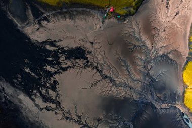 Üst taraftaki hava, İzlanda, Vik yakınlarında erimiş suyun karanlık volkanik kumu oyduğunu gösteriyor. Yosun sarısı yeşil kenarlar ve küçük kırmızı çatı yapısı yumuşak gün ışığında ölçek ekliyor..