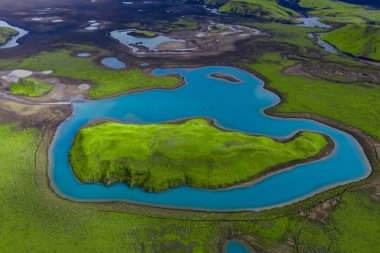 Havadan bakıldığında Landmannalaugar bölgesinde siyah kum, lav tarlaları, göletler ve hatta yaz gün ışığıyla birlikte yosun yeşil bir volkanik adanın etrafında turkuaz bir göl görülüyor..