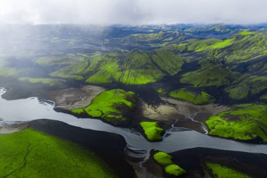 Havadan bakıldığında yosunlu volkanik tepeler ve İzlanda 'nın Landmannalaugar yakınlarındaki kara kum ovaları boyunca alçak bulutlar ve yumuşak yaz ışığı altında dönen bir buzul nehri görülüyor..