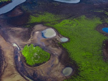 İzlanda volkanik dağlarının hava manzarası zümrüt yosun yatakları, koyu lav ve kül ovaları ve yumuşak ışık altında Landmannalaugar yakınlarındaki sütlü mavi jeotermal havuzları gösteriyor..