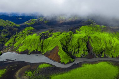 Hava görüntüsü, karanlık tepe ve vadilerin üzerinde neon yeşil yosunları, siyah kumların üzerinde örülmüş bir nehri ve Landmannalaugar yakınlarındaki İzlanda dağlarında alçak bulutları gösteriyor..
