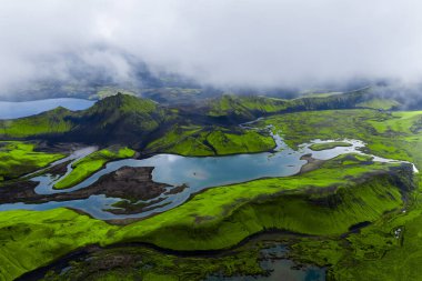 Alçak bulutlar, zümrüt yosunları ve karanlık lav düzlükleri Landmannalaugar yakınlarındaki İzlanda dağlık bölgelerinde yılan gölleriyle buluşurken serin yaz ışığı zıtlıkları yumuşatıyor..