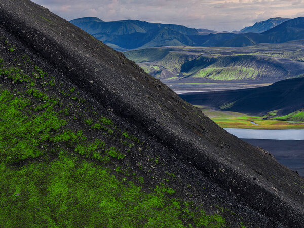 A diagonal ash ridge with neon moss cuts across volcanic terrain near Landmannalaugar, Iceland. Rhyolite hills and braided outwash flats extend under overcast light.