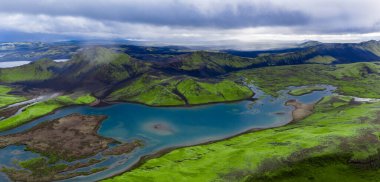 İzlanda 'da Landmannalaugar yakınlarındaki körfezlerde alçak bulutlar ve yumuşak gün ışığı altında yosun lav tarlaları ve karanlık kumların arasında geçen turkuaz bir göl manzarası..