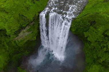 Hava görüntüsü Skogafoss şelalesinin sisli bir havuza düştüğünü gösteriyor, her iki tarafta yosunlu uçurumlar, yumuşak bulutlu ışık, doymuş yeşiller, İzlanda 'daki uzak volkanik arazi.