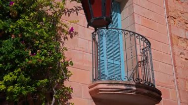 Güneş ışığıyla aydınlatılmış Mdina cephesinde süslü demir parmaklıklar, turkuaz kepenkler, mor bougainvillea ve kırmızı cam bir sokak feneri olan kavisli bir balkon görülüyor..