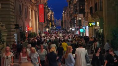 Valletta, Malta 'da kalabalık dar bir cadde müze pankartı, kireçtaşı cepheleri, balkonları ve alacakaranlık işaretleriyle kemere doğru ilerleyen bir bando gösteriyor..
