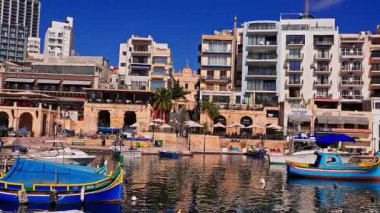 Sunlit Malta, Sliema limanı mavi ve sarı luzzu tekneleri, kireçtaşı kemerleri, kafeler ve Spinola Körfezi yakınlarında daireleri olan. İnsanlar yemek yer, gezer ve tekneler sakin sularda sallanır..