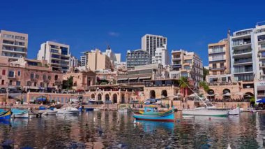 Malta, Sliema kasabasındaki panoramik öğlen manzarasında luzzu tekneleri ve yatları, kireçtaşı sütunları, kafeler ve modern bloklar görülüyor. Tekneler sallanır, su dalgalanır, insanlar rıhtımda hareket eder..