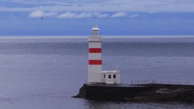 İzlanda 'da Keflavik yakınlarındaki bir deniz feneri sakin bir deniz kıyısındaki taştan bir dalgalanmadan yükselir, deniz kuşları daireler çizer ve tüneyerek, hafif dalgalanmalar sarar, sessiz tonlar çizgileri vurgular..