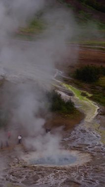 Yüksek açılı hava atışı İzlanda 'daki mavi bir havalandırmadan yükselen buharı gösteriyor. Turistler yakınlarda duruyor. Sarı yeşil rüzgarlar kahverengi zemin ve paslı kırmızı tepeler boyunca esiyor.