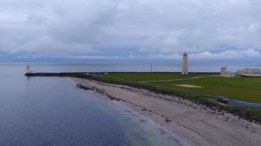 Hava görüntüsü, İzlanda kıyı şeridinde iki deniz feneri, servis binası ve araç gösteriyor. Kamera sahil boyunca ilerler ve sakin gri bir okyanusa doğru yol alır..
