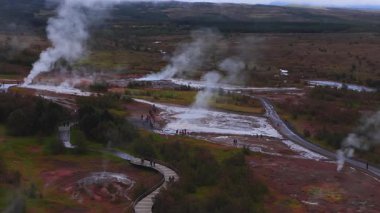 İzlanda 'daki Haukadalur' un hava manzarası buhar bulutları, fümeroleler, düz havzalar, dolambaçlı kaldırımlar, asfalt yollar ve bulutlu sonbahar ışığı altındaki ziyaretçileri gösteriyor..