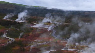 İzlanda 'daki Haukadalur Geysir' in hava manzarasında dumanı tüten fümeroller, sıcak havuzlar, merkezi bir havuz, tahta kaldırımlarda ve patikalarda ziyaretçiler ve mineral toprakları üzerindeki bulutlu ışık görülüyor..