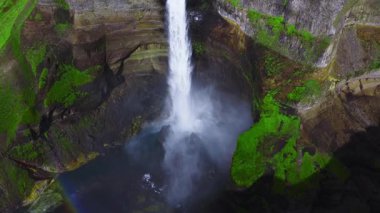 Hava görüntüsü Haifoss 'un İzlanda, Thjorsardalur' da oyulmuş bir amfi tiyatroya daldığını gösteriyor. Parlak gün ışığı, katmanlı volkanik kayayı, zümrüt yosunu, sisi ve soluk bir gökkuşağını ortaya çıkarır..