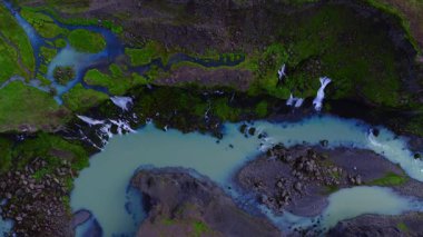 Yukarıdan bakıldığında, İzlanda 'da küçük şelaleleri ve koyu bazalt kayalıkları olan yosun kaplı bir volkanik kanyonda örülmüş sütlü bir turkuaz buzul nehri görülüyor..