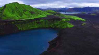Havadan bakıldığında İzlanda, Landmannalaugar yakınlarında kobalt krater gölü, yosun kaplı jantlar ve siyah kül ovaları görülüyor. Yüksek doygunluk ile bulutlu gün ışığında yavaş süzül.