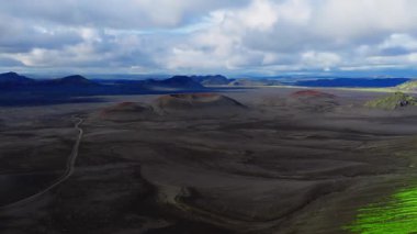 Hava görüntüsü, İzlanda 'da siyah kül tarlasında paslı kırmızı jantlı kül konileri, dolambaçlı toprak bir yol, mavi dağlar, parlak yeşil yosun ve yavaş çekim..