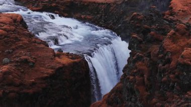 Hava görüntüsü, Thjorsardalur, İzlanda 'da Haifoss' un turkuaz akıntıların dar bir kanyona düştüğünü, sisin yükseldiğini ve bazalt duvarların ve turuncu yosunların sahneyi tanımladığını gösteriyor..