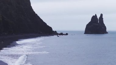 Reynisfjara 'nın kara kumları, bazalt kayalıkları ve İzlanda, Vik yakınlarındaki Reynisdrangar' ın hava sahili manzarası. Küçük gruplar dalgalar gibi yürür, serin mavi gri tonlar, bulutlu.