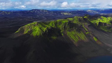 Bir hava aracı İzlanda volkanik ovaları üzerinde süzülür, krater sırtlarında yosun, sağ altta küçük bir krater gölü, alçak bulutlar ve yumuşak gün ışığı dokusu ve ölçeği ortaya çıkarır..