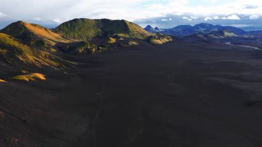 İzlanda dağlarının hava aracı görüntüsü siyah kum düzlükleri, yosunlu ryolit dağları, kraterler, nehir kıvrımları, 4x4 pist ve sıcak, düşük ışıkta park edilmiş bir araç gösteriyor..