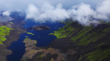 Hava görüntüsü volkanik kobalt mavi krater gölü, siyah lav kumları, yeşil yosun tepeleri ve yumuşak bulutlu ışık altında İzlanda dağlarında sürüklenen sisi gösteriyor..