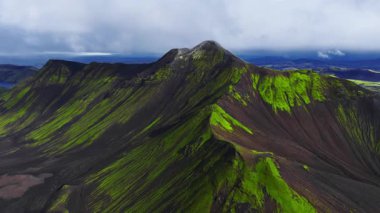 Landmannalaugar yakınlarındaki İzlanda Dağları 'ndaki kraterli bir volkanik dağın üzerinden geçen hava kamerası neon yeşil yosun, koyu kül yamaçları ve sürüklenen sisli....