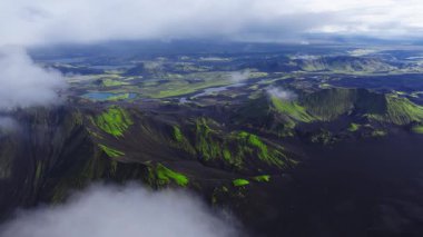 İzlanda Dağları 'nın yosun yeşil kraterleri, turkuaz gölleri ve yumuşak bulutlu ışık altında örülmüş nehirleriyle hava manzarası. Yavaş sinematik hareket sırtları ortaya çıkarır.