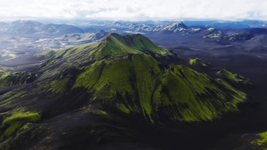 Havadan bakıldığında İzlanda dağlarında yosun kaplı külahlar, kraterli tepeler ve siyah kül ovaları görülüyor. Geniş çerçeveli yumuşak gün ışığında kamera izleri ve tavalar.