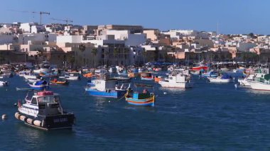 Hava görüntüsü Malta 'daki Marsaxlokk limanını, luzzu' yu ve çalışma teknelerini gösteriyor. Mavi suda sallanıyor, palmiyeler ve tentelerle aydınlatılmış kireçtaşı iskelede, vinçlerin arkasında yeni bloklar var..
