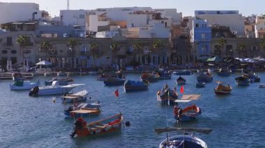 Marsaxlokk 'un hava görüntüsünde, Malta palmiye astarlı iskele, taş rıhtım, pastel evler, kafeler, bayraklar, şamandıralar ve öğleden sonra ışıklarının yanında sallanan luzzu tekneleri görülüyor..