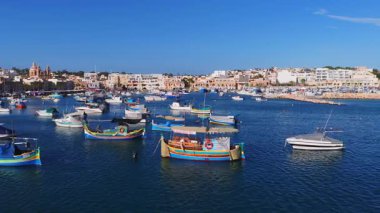 Malta 'daki Marsaxlokk limanının hava görüntüsünde, öğle vakti mavi su, kum renkli binalar, kubbeli kilise, dalgakıran ve marina üzerinde luzzu botları ve zanaatı görülüyor..