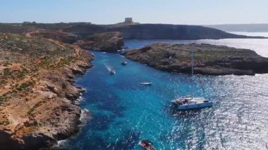 St. Marys Kulesi, yatlar ve katamaran hareketleriyle Malta 'daki Comino Kanalı' nın hava manzarası. İnsanlar gün ortasında güçlü bir ışık altında yüzerken berrak sularda ilerliyorlar..