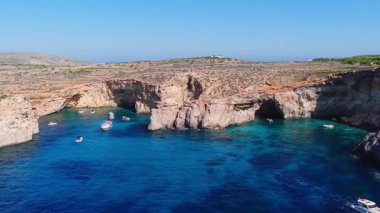 Hava panoramik görüntüsü, Malta 'nın Comino Adası' nda kireçtaşı kayalıkları, deniz mağaraları, bir kaya kemeri, turkuaz körfez, yat ve tekneler, daireler çizen yüzücüler ve gün ortası ışığı olduğunu gösteriyor..