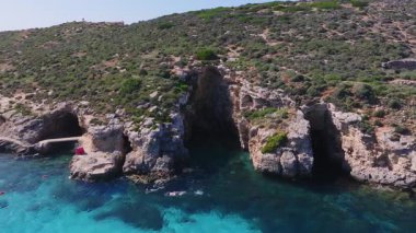 Kireçtaşı kayalıkları, deniz mağaraları, beton iskelesi, çakıl taşı plajı, işaret şamandıraları, turistler ve gün ortasında yüzen yüzücülerle Malta 'daki Comino Adası' nın hava manzarası..