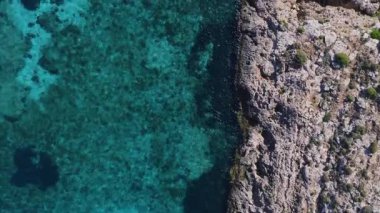 Malta 'daki Comino Adası' nın havadan aşağı manzarası. Sağ engebeli kireçtaşı kıyı şeridi ve açık turkuaz deniz. Su altı kayaları ve karanlık havuzlar renk bozulmalarını gösterir.